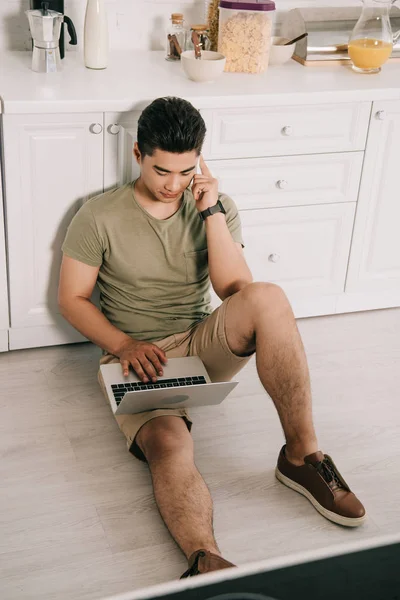 Vista ad alto angolo di giovane uomo asiatico parlando su smartphone e utilizzando il computer portatile mentre seduto sul pavimento in cucina — Foto stock