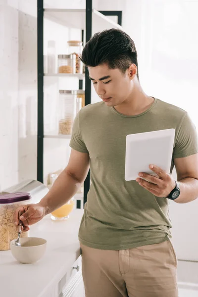 Bonito ásia homem mistura flocos no tigela enquanto segurando digital tablet no cozinha — Fotografia de Stock