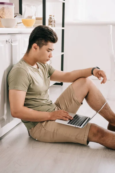 Attentive asian man using laptop while sitting on floor in kitchen — Stock Photo