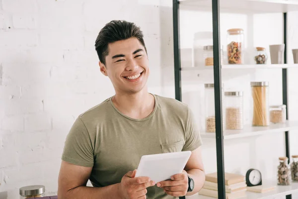 Cheerful asian man holding digital tablet while smiling at camera in kitchen — Stock Photo