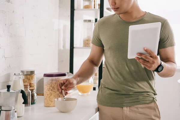 Vista parziale del giovane che mescola i fiocchi nella ciotola mentre tiene la tavoletta digitale in cucina — Foto stock