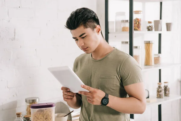 Concentrado asiático hombre usando digital tablet mientras de pie cerca cocina mesa - foto de stock