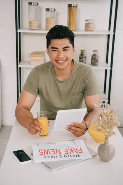 Fröhlicher asiatischer Mann mit digitalem Tablet und Glas Orangensaft, während er am Küchentisch neben Fake News Zeitung und Smartphone mit leerem Bildschirm sitzt — Stockfoto