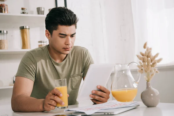 Bello asiatico uomo guardando digitale tablet mentre seduto a cucina tavolo e tenendo bicchiere di arancia succo — Foto stock