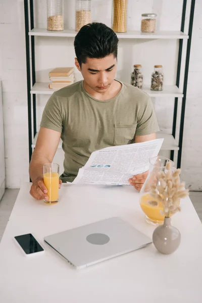 Внимательный азиатский мужчина читает газету, сидя за кухонным столом и держа стакан апельсинового сока — стоковое фото