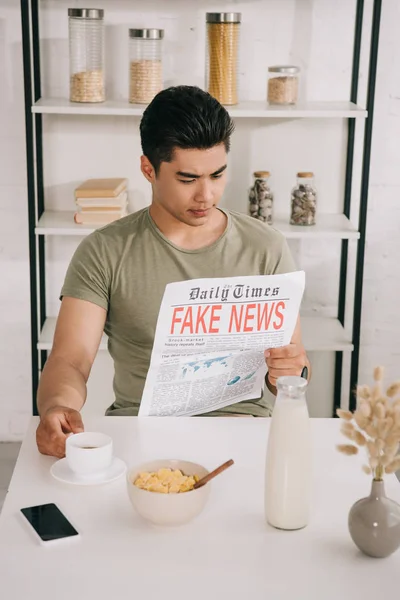 Bonito ásia homem leitura jornal com falso notícias enquanto sentado no cozinha mesa perto de xícara de café, tigela com flocos e garrafa de leite — Fotografia de Stock