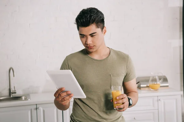 Young asian man using digital tablet while standing in kitchen with glass of orange juice — Stock Photo