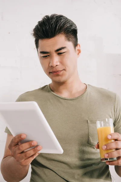 Guapo asiático hombre leyendo falso noticias periódico mientras celebración de vaso de naranja jugo - foto de stock