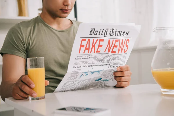 Ausschnitt eines jungen Mannes, der Fake News Zeitung liest, während er am Küchentisch sitzt und ein Glas Orangensaft in der Hand hält — Stockfoto