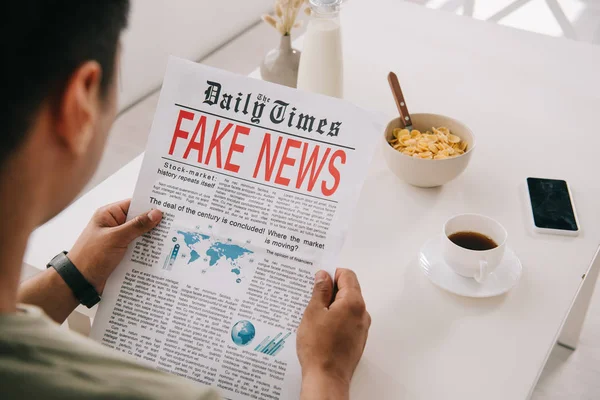 Cropped view of man reading newspaper with fake news while sitting at kitchen table near cup of coffee, bowl with flakes and smartphone with blank screen — Stock Photo