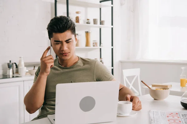 Ernster asiatischer Mann spricht auf Smartphone, während er am Küchentisch sitzt und Laptop benutzt — Stockfoto