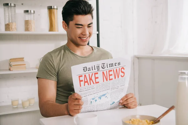 Молодой азиат улыбается, сидя за кухонным столом и читая фейковую газету — стоковое фото