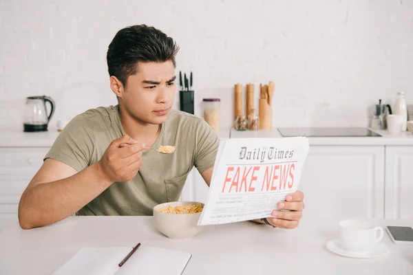 Bello asiatico uomo avendo colazione mentre lettura falso news giornale in cucina — Foto stock