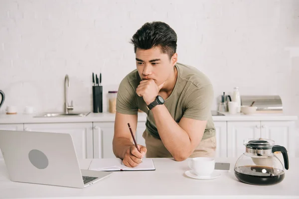 Jeune asiatique homme holiding crayon tout en regardant ordinateur portable dans la cuisine — Photo de stock