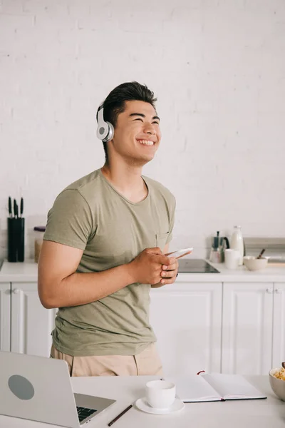 Alegre ásia homem ouvindo música no hedphones enquanto segurando smartphone e olhando para longe — Fotografia de Stock