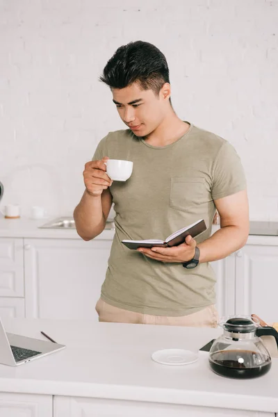 Junger asiatischer Mann trinkt Kaffee, während er Laptop und Notizbuch in der Hand hält — Stockfoto
