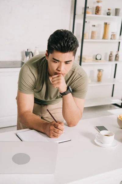 Attentive asian man writing notebook while looking at laptop in kitchen — Stock Photo