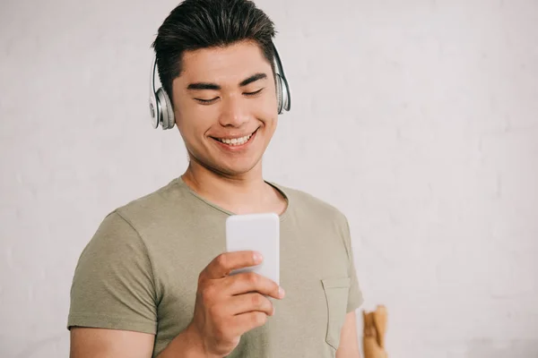 Веселый азиатский мужчина слушает музыку в наушниках, глядя на смартфон — стоковое фото