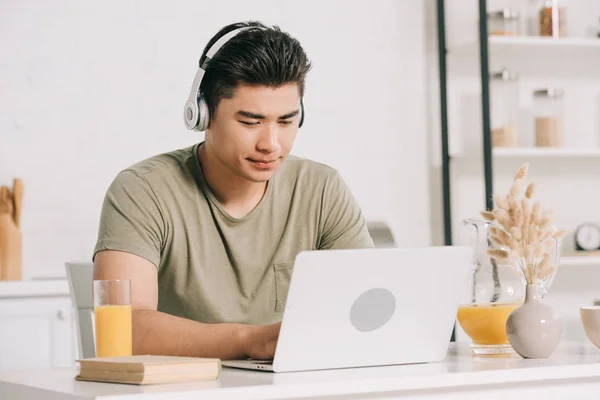 Concentrado asiático homem em fones de ouvido usando laptop enquanto sentado na mesa da cozinha — Fotografia de Stock