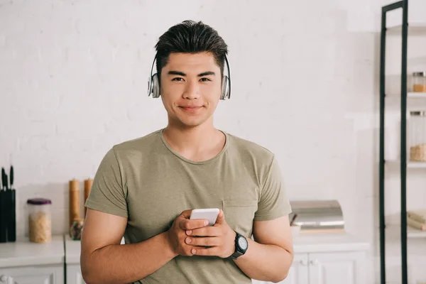 Schöner asiatischer Mann mit Kopfhörern lächelt in die Kamera, während er sein Smartphone in der Hand hält — Stockfoto