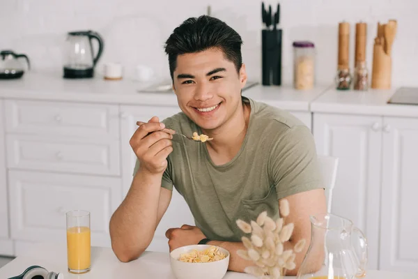 Junger asiatischer Mann frühstückt, während er am Küchentisch sitzt und in die Kamera lächelt — Stockfoto