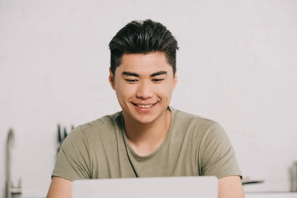 Веселый азиатский человек с помощью ноутбука, сидя за кухонным столом — стоковое фото