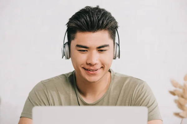 Alegre asiático hombre usando laptop y escuchar música en auriculares - foto de stock