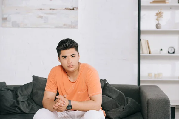 Nachdenklicher asiatischer Mann schaut in die Kamera, während er zu Hause auf dem Sofa sitzt — Stockfoto