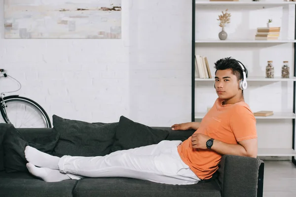 Задумливий азіатський чоловік в навушниках дивиться на камеру, відпочиваючи на дивані вдома — стокове фото