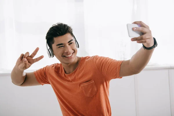 Happy asian man taking selfie while showing victory gesture and listening music in headphones — Stock Photo