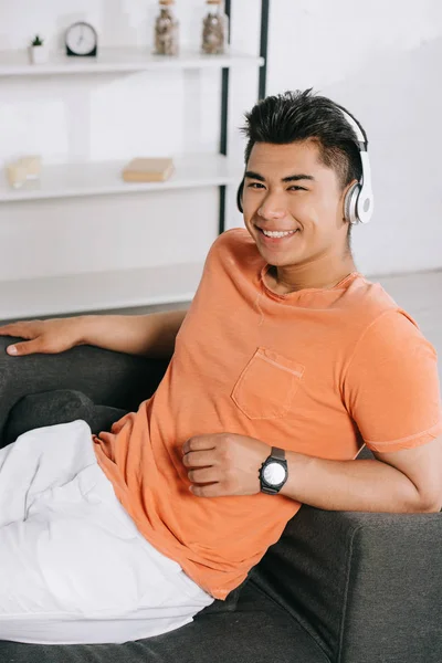 Красивый азиатский мужчина улыбается в камеру, отдыхая на диване и слушая музыку в наушниках — стоковое фото