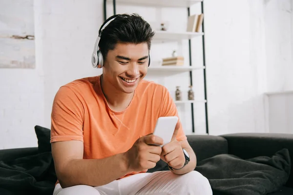 Cheerful asian man using smartphone and listening music in headphones while sitting on sofa at home — Stock Photo