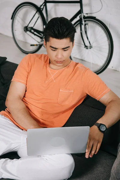 Concentrated asuan man using laptop while sitting on sofa at home — Stock Photo