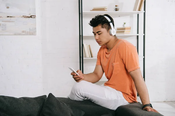Jovem asiático homem ouvindo música em fones de ouvido e usando smartphone em casa — Fotografia de Stock