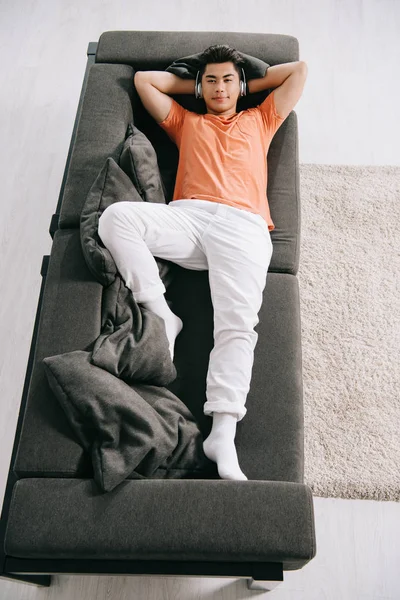 Von oben: junger asiatischer Mann hört Musik über Kopfhörer, während er zu Hause auf dem Sofa liegt — Stockfoto