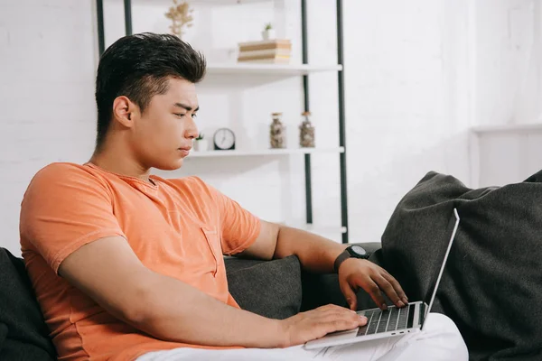 Junger, aufmerksamer asiatischer Mann sitzt zu Hause auf der Couch mit Laptop — Stockfoto