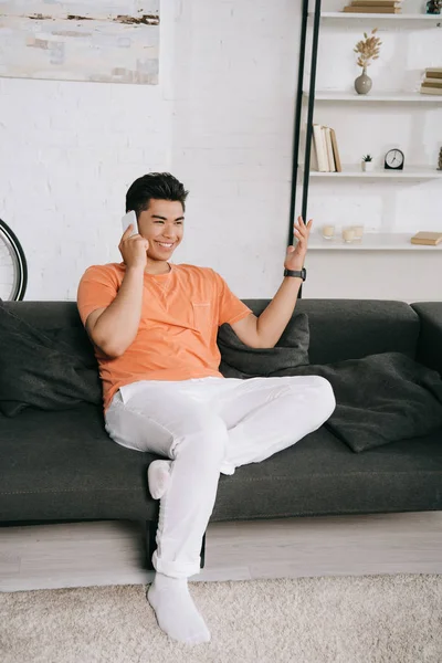 Cheerful asian man gesturing while sitting on couch and talking on smartphone — Stock Photo