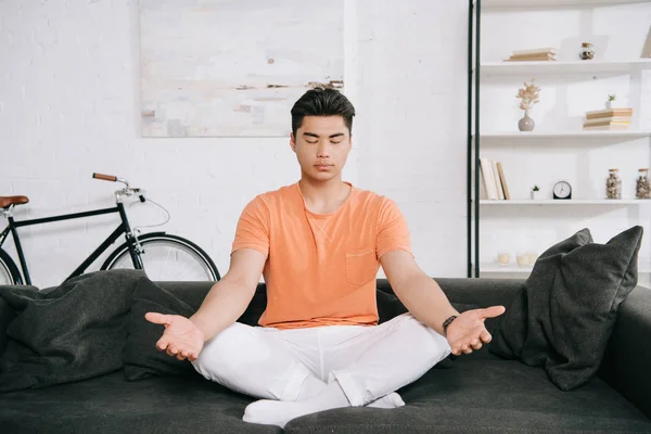 Jovem asiático homem meditando enquanto sentado em lótus posar no sofá em casa — Fotografia de Stock