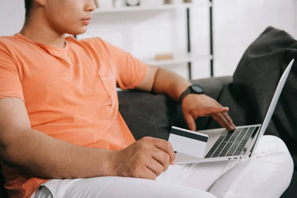 Vista ritagliata del giovane utilizzando il computer portatile e tenendo la carta di credito mentre seduto sul divano a casa — Foto stock