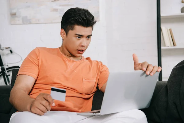 Surprised asian man looking at laptop while sitting on couch and holding credit card — Stock Photo