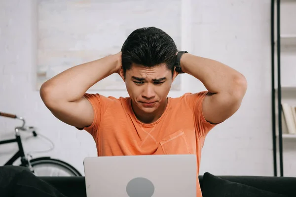 Scosso asiatico uomo holding mani dietro testa mentre seduta su divano e guardando laptop — Foto stock