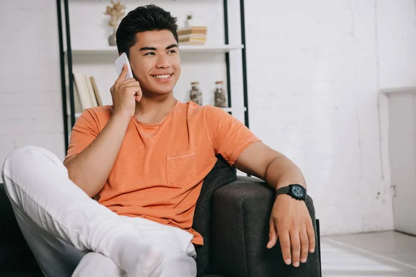 Fröhlicher asiatischer Mann schaut weg, während er auf dem Sofa sitzt und auf dem Smartphone spricht — Stockfoto