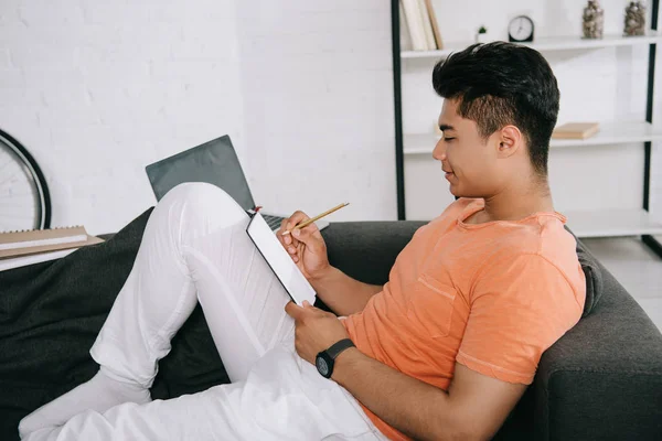 Young asian man writing in notebook while sitting on couch at home — Stock Photo