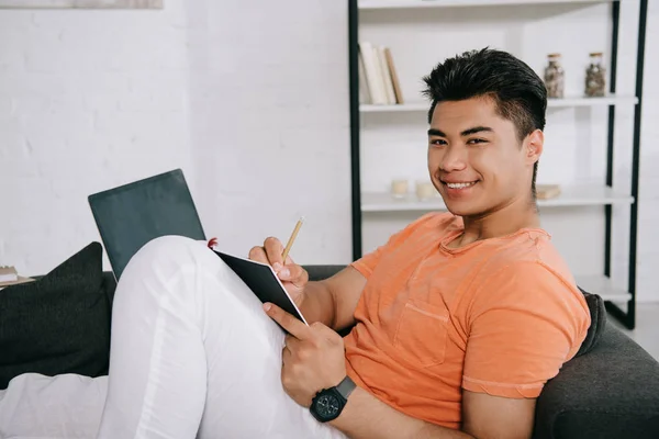 Handsome asian man smiling at camera while lying on sofa and writing in notebook — Stock Photo