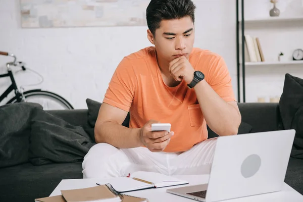 Premuroso asiatico uomo guardando laptop mentre seduto sul divano e tenendo smartphone — Foto stock