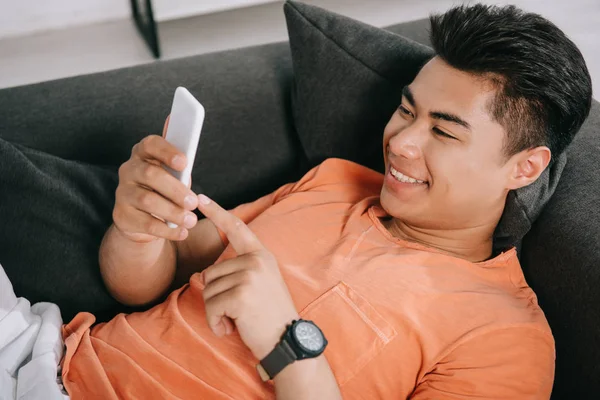 Alegre asiático homem usando smartphone enquanto relaxa no sofá em casa — Fotografia de Stock