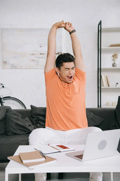 Tired asian man stretching and yawning while sitting on sofa near table with laptop and notebooks — Stock Photo