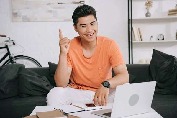 Alegre asiático hombre mirando cámara y mostrando idea signo mientras sentado en escritorio con portátil - foto de stock