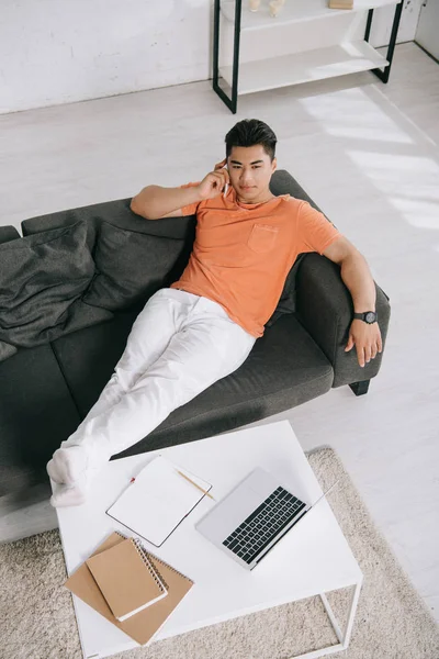 High angle view of handsome asian man talking on smartphone while lying on sofa near desk with laptop — Stock Photo