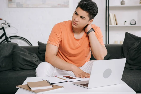 Cansado asiático hombre tocando cuello mientras sentado cerca de mesa con laptop y notebooks - foto de stock